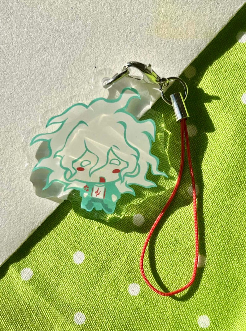 May include: A clear acrylic charm featuring a cartoon character with white hair and teal outlines. The charm has a red lanyard and a silver clasp. The character has a teal outfit and a red face. The charm is on a green and white polka dot background.