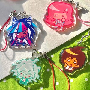 May include: A collection of colorful acrylic charms featuring anime-style character designs. Each charm has a unique character with vibrant colors, attached to a metal clasp and a colored lanyard. The charms are displayed on a white and green polka dot background.