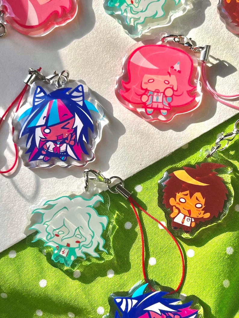 May include: A collection of colorful acrylic charms featuring anime-style character designs. Each charm has a unique character with vibrant colors, attached to a metal clasp and a colored lanyard. The charms are displayed on a white and green polka dot background.