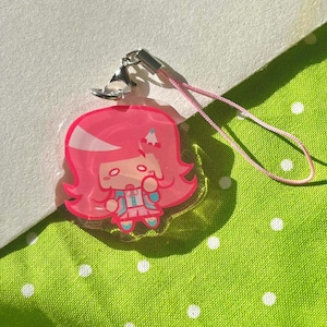 May include: A pink acrylic charm featuring a cartoon character with pink hair and a blue outfit. The charm has a silver clasp and a pink lanyard. The background is a green fabric with white polka dots.