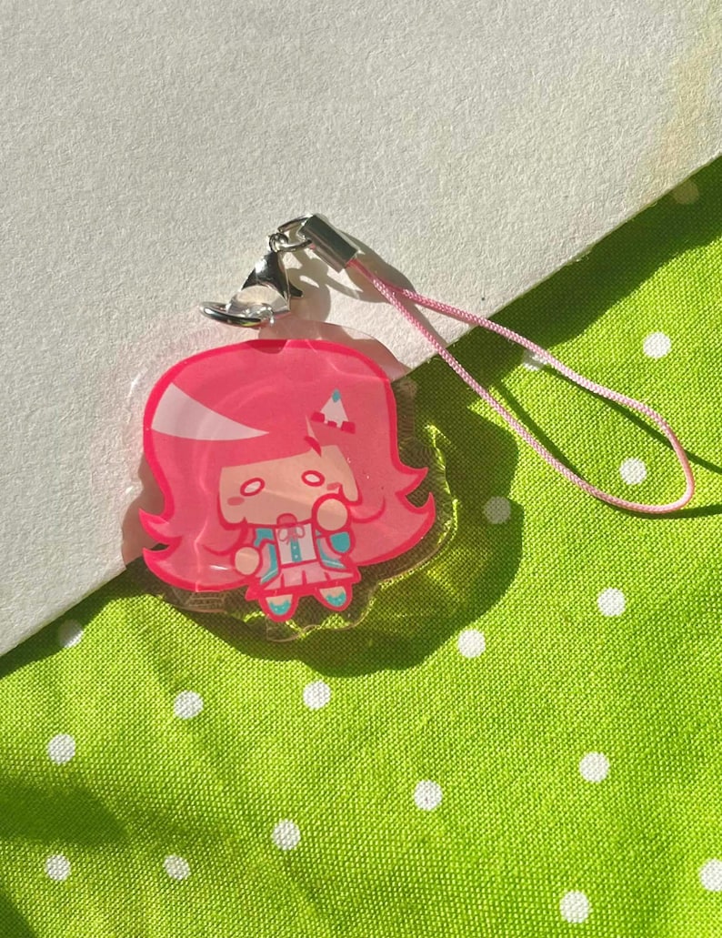 May include: A pink acrylic charm featuring a cartoon character with pink hair and a blue outfit. The charm has a silver clasp and a pink lanyard. The background is a green fabric with white polka dots.
