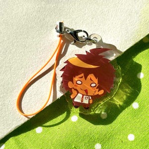 May include: A clear acrylic charm featuring a cartoon character with spiky brown hair and a white shirt. The charm has an orange lanyard and a silver clasp. The background is a light green fabric with white polka dots.