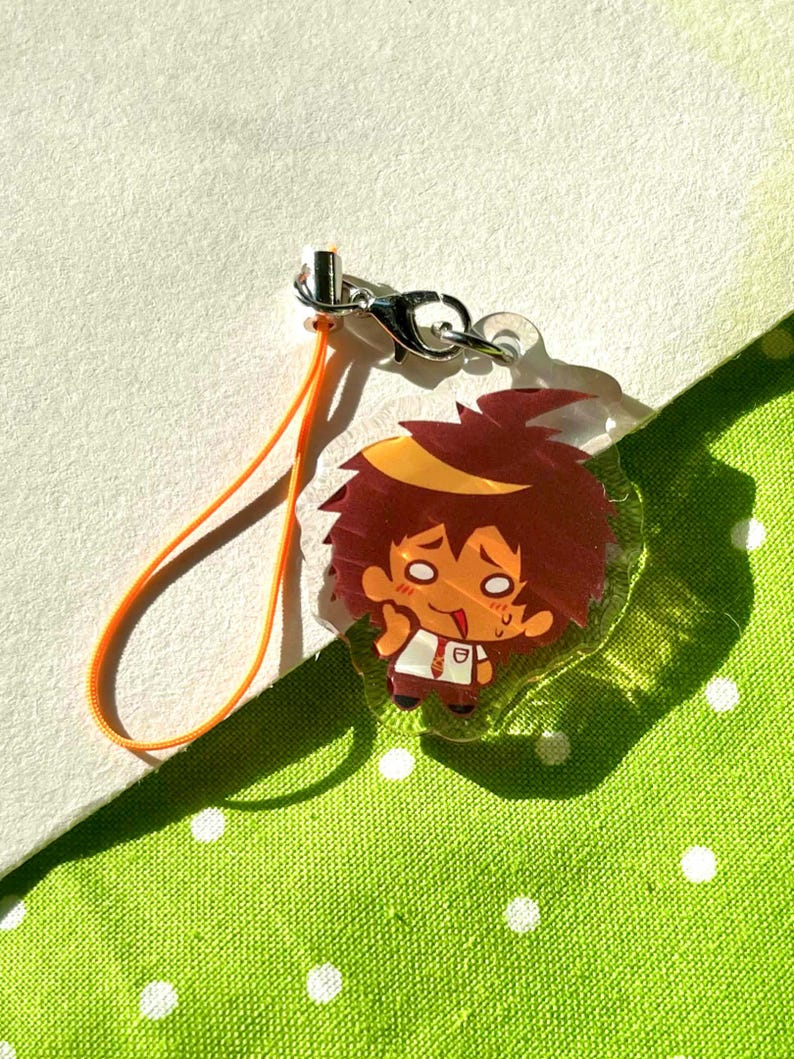 May include: A clear acrylic charm featuring a cartoon character with spiky brown hair and a white shirt. The charm has an orange lanyard and a silver clasp. The background is a light green fabric with white polka dots.