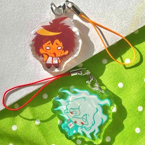 May include: Two anime-style acrylic charms with silver-tone clasps and colored cords. One charm features a red-haired character with a yellow headband, the other a white-haired character with teal accents. The background is a green polka-dot fabric.