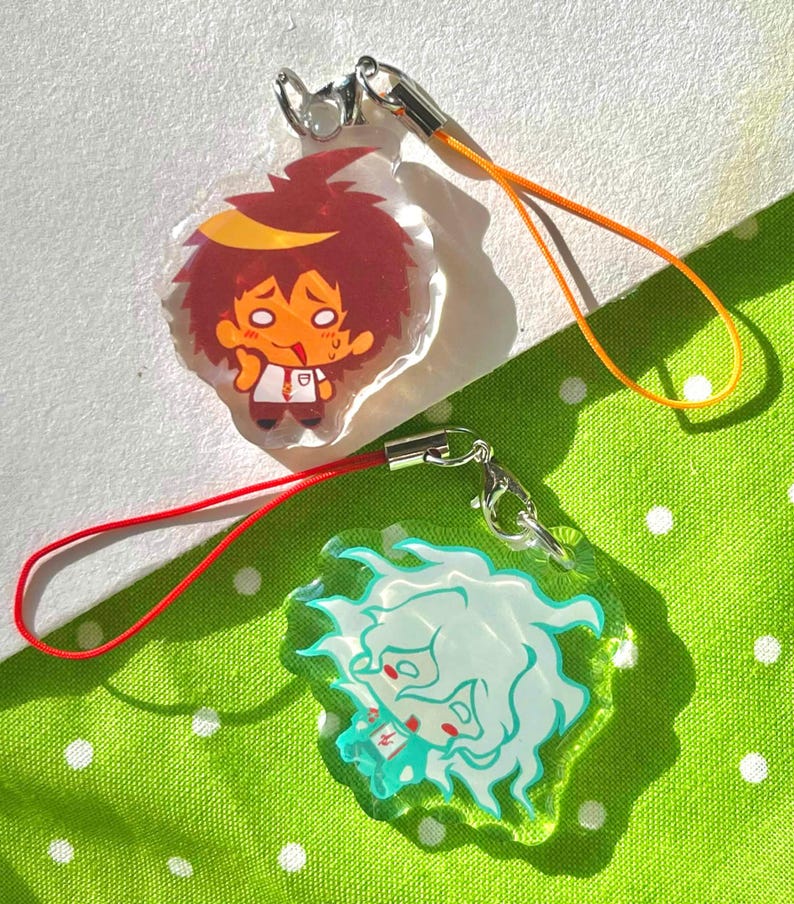 May include: Two anime-style acrylic charms with silver-tone clasps and colored cords. One charm features a red-haired character with a yellow headband, the other a white-haired character with teal accents. The background is a green polka-dot fabric.