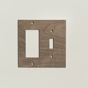 May include: A wooden light switch cover with a rectangular opening for a rocker switch and a smaller opening for a toggle switch. The cover is a light brown color and has a square shape with screw holes.