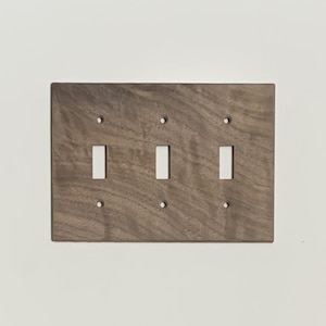 May include: A wooden triple light switch plate with a dark brown finish. The plate has three rectangular openings for the switches and four screw holes. The wood grain is visible.