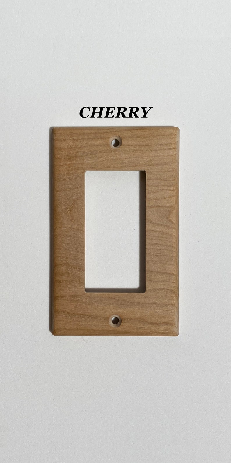 May include: A light brown wooden wall plate with two holes for mounting. The plate has a rectangular cutout for a light switch or outlet. The text "CHERRY" is printed above the plate.