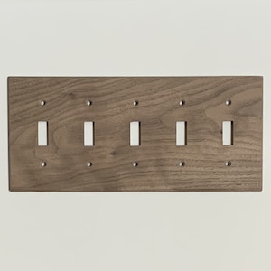 May include: A wooden wall plate with six cutouts for light switches. The plate is a dark brown color and has a wood grain pattern.