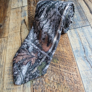 May include: A necktie featuring a realistic forest camouflage pattern with shades of black, gray, and white, accented by detailed brown and green oak leaf prints. The tie is rolled at the top and tapers to a pointed end at the bottom.