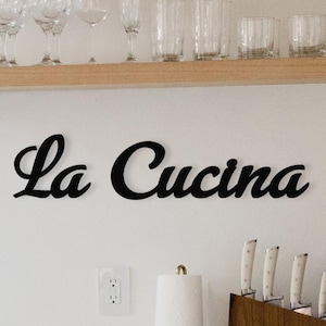 May include: A black wooden sign with the words "La Cucina" in a cursive font. The sign is mounted on a white wall in a kitchen. There are glasses on a shelf above the sign, and a coffee maker and a knife block on the counter below.