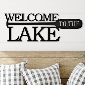 May include: Black metal wall sign with the words "Welcome to the Lake" and a stylized paddle design.