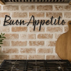 May include: Black metal "Buon Appetito" sign mounted on a brick wall above a gas stove. A wooden cutting board and a potted plant are also visible. The kitchen decor has a rustic, farmhouse style.