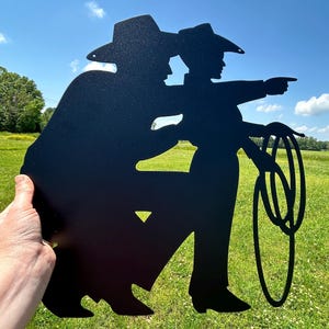 Puede incluir: Silueta met&aacute;lica negra de dos vaqueros, uno se&ntilde;alando, el otro sosteniendo un lazo. La obra de arte se sostiene sobre un fondo de cielo azul brillante y hierba verde.