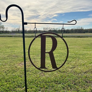 May include: Black metal garden flag stand with a round brown metal sign featuring the letter "R" in a circle.
