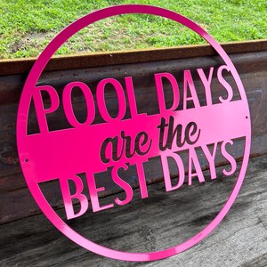 May include: A bright pink metal sign with the words "POOL DAYS are the BEST DAYS" cut out. The sign is circular with a horizontal bar across the center. The sign is displayed on a weathered wooden surface.