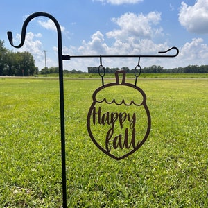 May include: A metal garden stake with a decorative acorn-shaped sign that reads "Happy Fall" in a cursive font. The sign is brown and hangs from a black metal hook. The background is a grassy field under a blue sky.