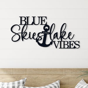 A black metal sign with the text 'BLUE SKIES AND LAKE VIBES' with an anchor symbol in between the words 'SKIES' and 'LAKE'. The sign is mounted on a white wall above a wooden bench with two checkered pillows.