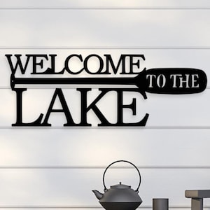 May include: Black metal wall sign with the text "Welcome to the Lake" with a stylized paddle design.