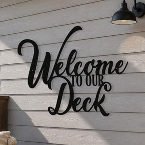 May include: Black metal sign that reads "Welcome to Our Deck" in a decorative script font. The sign is mounted on a light gray, wood-paneled wall, with a black outdoor light fixture above. A chair with a striped pillow is visible.