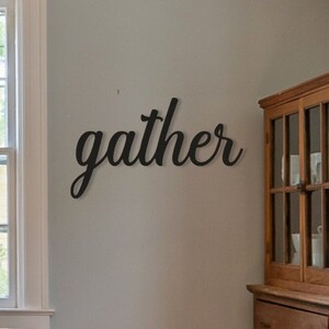 May include: Black metal wall decor with the word "gather" in a cursive font. The decor is mounted on a light gray wall in a dining room setting.