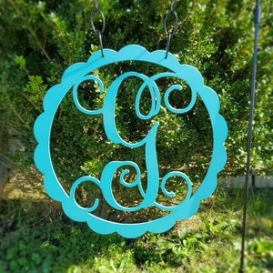 May include: A teal blue wooden sign with a scalloped edge and a decorative letter G in a cursive font. The sign is hanging from a black metal stake.