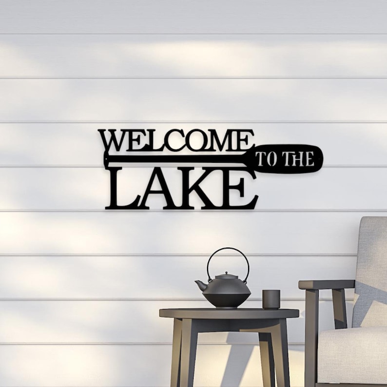 May include: Black metal wall sign with the text "Welcome to the Lake" with a stylized paddle design.
