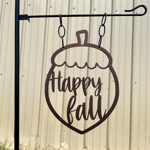 May include: A brown metal garden flag shaped like an acorn, hanging from a black metal pole. The words "Happy Fall" are cut out within the acorn shape. The flag is suspended by chains.