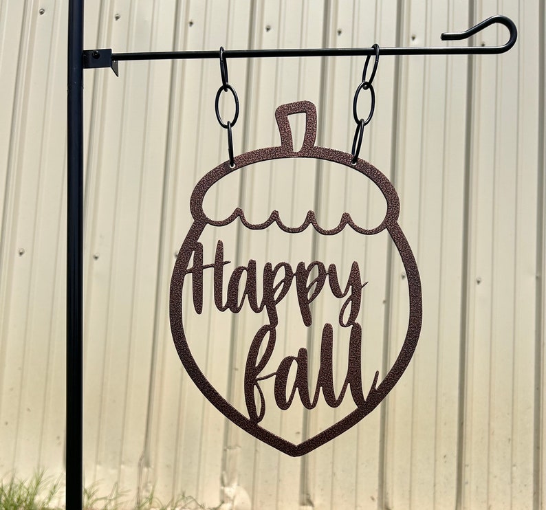 May include: A brown metal garden flag shaped like an acorn, hanging from a black metal pole. The words "Happy Fall" are cut out within the acorn shape. The flag is suspended by chains.
