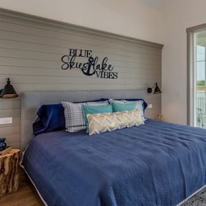 May include: A blue and gray bedroom with a bed with blue bedding and pillows. The headboard has a wooden sign that reads "Blue Skies & Lake Vibes".