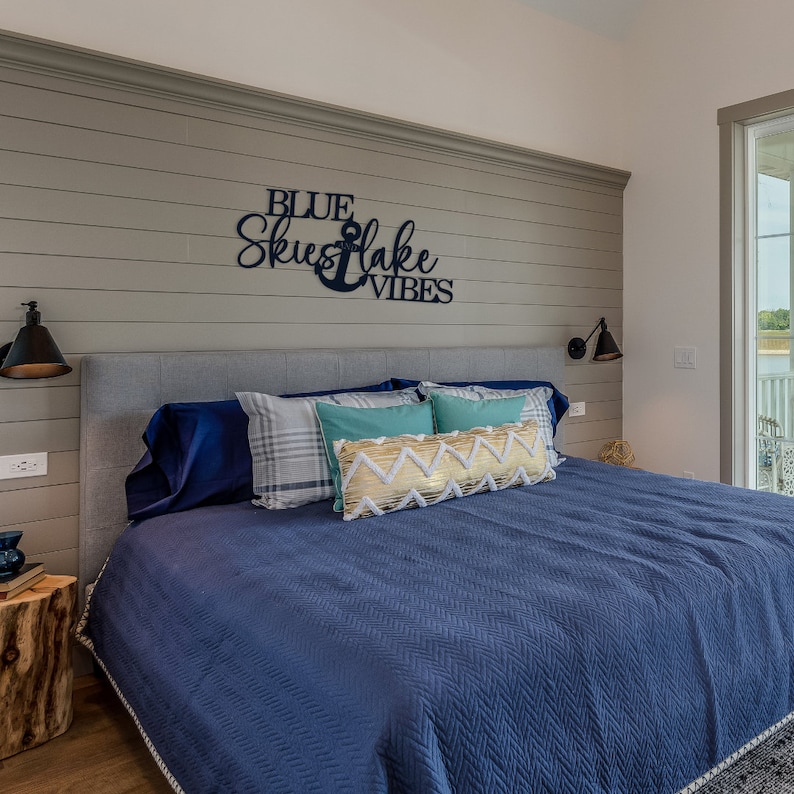 May include: A blue and gray bedroom with a bed with blue bedding and pillows. The headboard has a wooden sign that reads "Blue Skies & Lake Vibes".