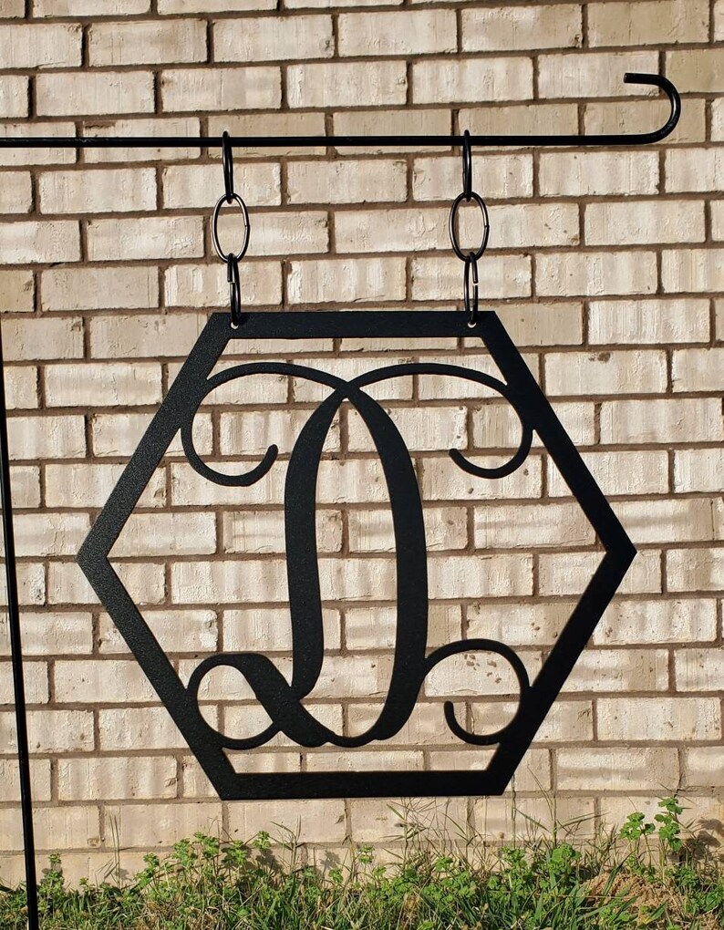 K&ouml;nnte beinhalten: Schwarzes sechseckiges Schild aus Metall mit einem dekorativen Buchstaben D in der Mitte. Das Schild h&auml;ngt an einer schwarzen Metallstange mit zwei Ketten.