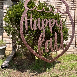 May include: A bronze-colored metal sign in the shape of a pumpkin with the words "Happy Fall" in a cursive font. The sign is hanging in front of a green bush and a brick wall. The sign is suspended by a black chain.