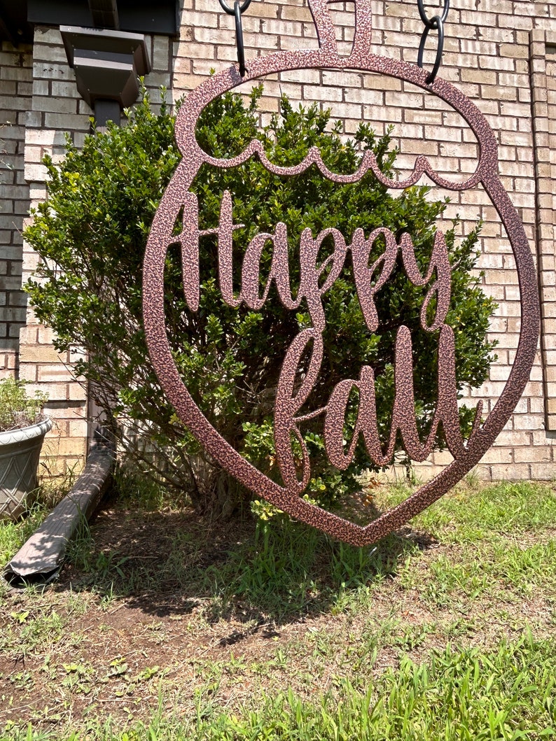 May include: A bronze-colored metal sign in the shape of a pumpkin with the words "Happy Fall" in a cursive font. The sign is hanging in front of a green bush and a brick wall. The sign is suspended by a black chain.