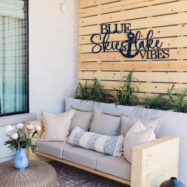 A light gray couch with three cushions and a long throw pillow. The couch is made of wood and has a light wood frame. The couch is in front of a wooden wall with a blue metal sign that reads 'Blue Skies and Lake Vibes' with an anchor symbol. There are green plants in a white planter in front of the wall.
