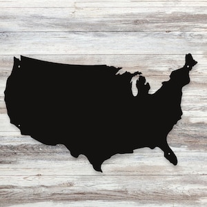 May include: A black metal cutout of the United States of America, mounted on a light-colored, weathered wooden background. The cutout is a solid silhouette of the country's shape, with visible mounting holes. The wood grain adds a rustic touch.