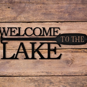 May include: Black metal sign with the words "Welcome to the Lake" and a stylized paddle design.