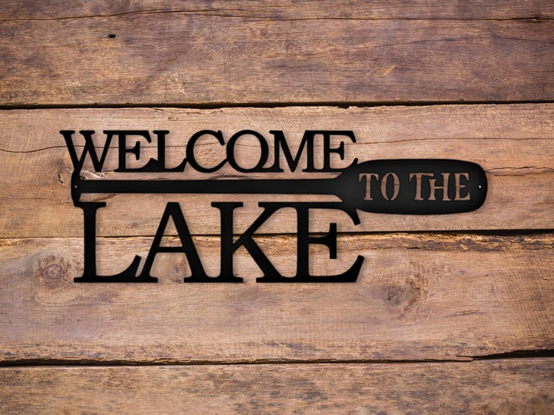 May include: Black metal sign with the words "Welcome to the Lake" and a stylized paddle design.