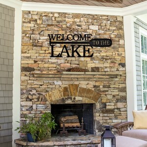 May include: Black metal sign with the words "Welcome to the Lake" on a wooden paddle shape. The sign is mounted on a stone fireplace.