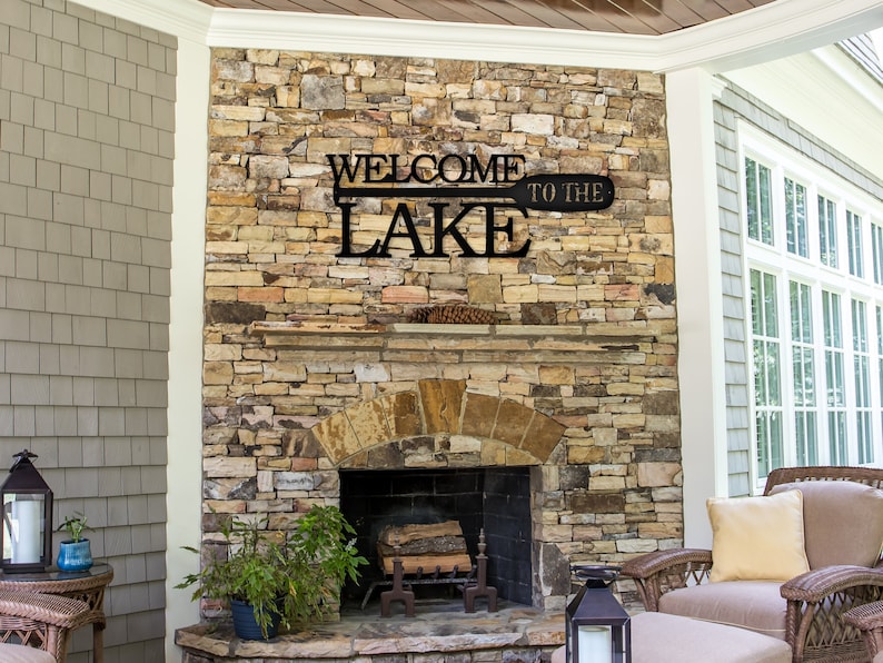 May include: Black metal sign with the words "Welcome to the Lake" on a wooden paddle shape. The sign is mounted on a stone fireplace.
