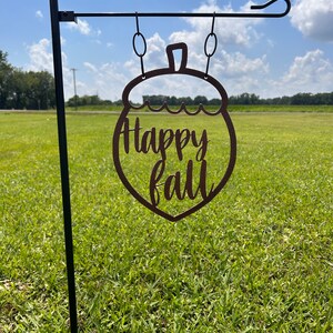 May include: A brown metal garden flag shaped like an acorn, with the words "Happy fall" cut out. The flag hangs from a black metal pole, set against a backdrop of green grass and a blue sky.