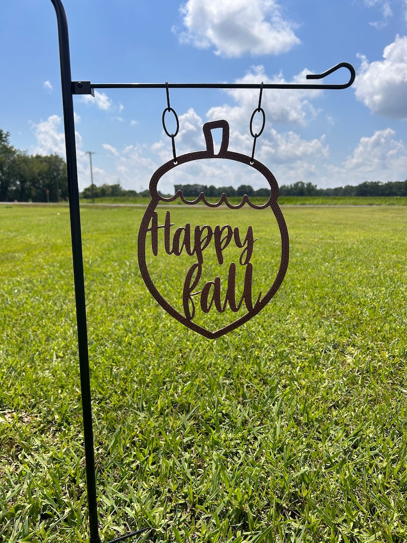 May include: A brown metal garden flag shaped like an acorn, with the words "Happy fall" cut out. The flag hangs from a black metal pole, set against a backdrop of green grass and a blue sky.