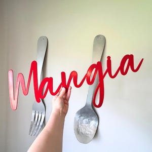May include: Red metal wall art with the word "Mangia" in a cursive font. The art is hanging on a white wall with two large silver utensils behind it.