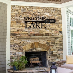 May include: Black metal sign with the words "Welcome to the Lake" on a wooden paddle design. The sign is mounted on a stone fireplace in a porch setting.