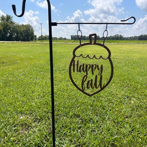 May include: A metal garden stake with a decorative acorn-shaped sign that reads "Happy Fall" in a cursive font. The sign is brown and hangs from a black metal pole, set against a green field and a blue sky.