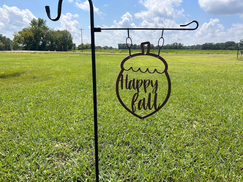 May include: A metal garden stake with a decorative acorn-shaped sign that reads "Happy Fall" in a cursive font. The sign is brown and hangs from a black metal pole, set against a green field and a blue sky.