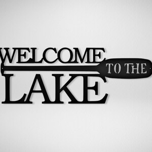 May include: Black metal sign with the words "Welcome to the Lake" and a silhouette of a paddle.