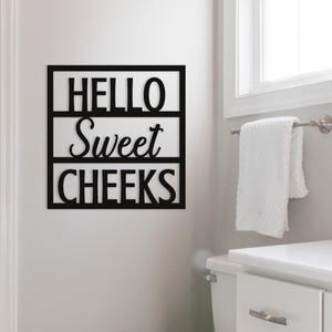 May include: Black wooden wall art with the words "HELLO Sweet CHEEKS" in a bathroom setting. The sign is rectangular with the text arranged in three lines. A white towel hangs on a chrome towel rack near a window.