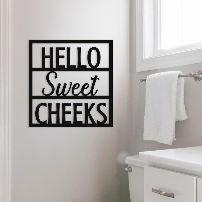 May include: Black wooden wall art with the words "HELLO Sweet CHEEKS" in a bathroom setting. The sign is rectangular with the text arranged in three lines. A white towel hangs on a chrome towel rack near a window.