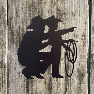 Puede incluir: Silueta met&aacute;lica negra de dos vaqueros, uno arrodillado y el otro se&ntilde;alando, con un lazo. La obra de arte est&aacute; montada sobre un fondo de madera envejecida, evocando un tema r&uacute;stico y del Oeste.